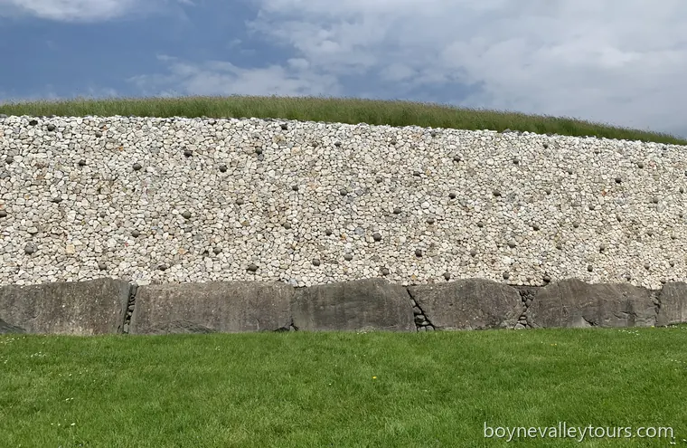 White quartz and dark river-stone facade of Newgrange above grey kerbstones under a blue sky.