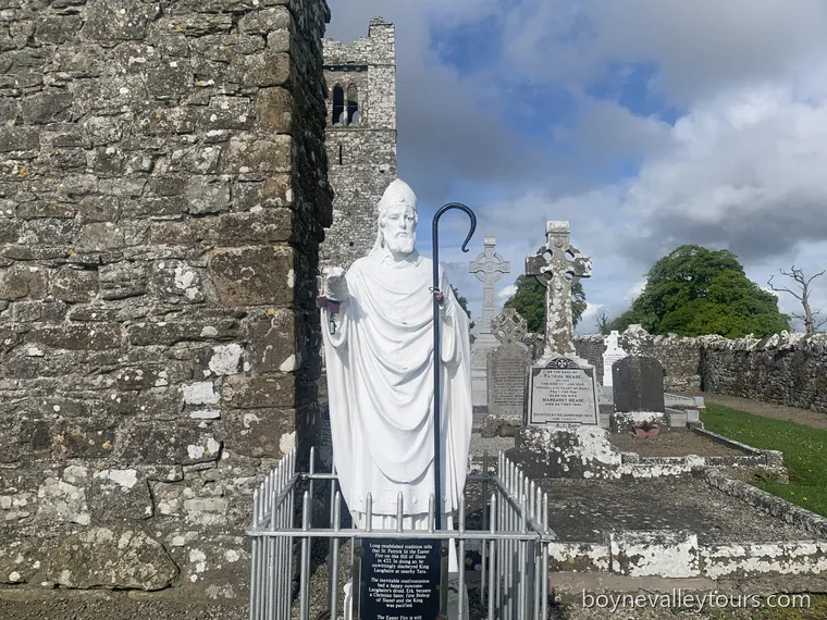 White statue of Saint Patrick in the Hill of Slane graveyard, with the friary tower and Celtic crosses behind.